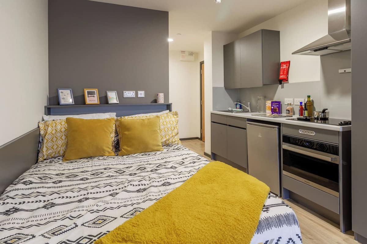 A compact room with a bed, a kitchenette, and a grey wall with a shelf holding framed pictures.