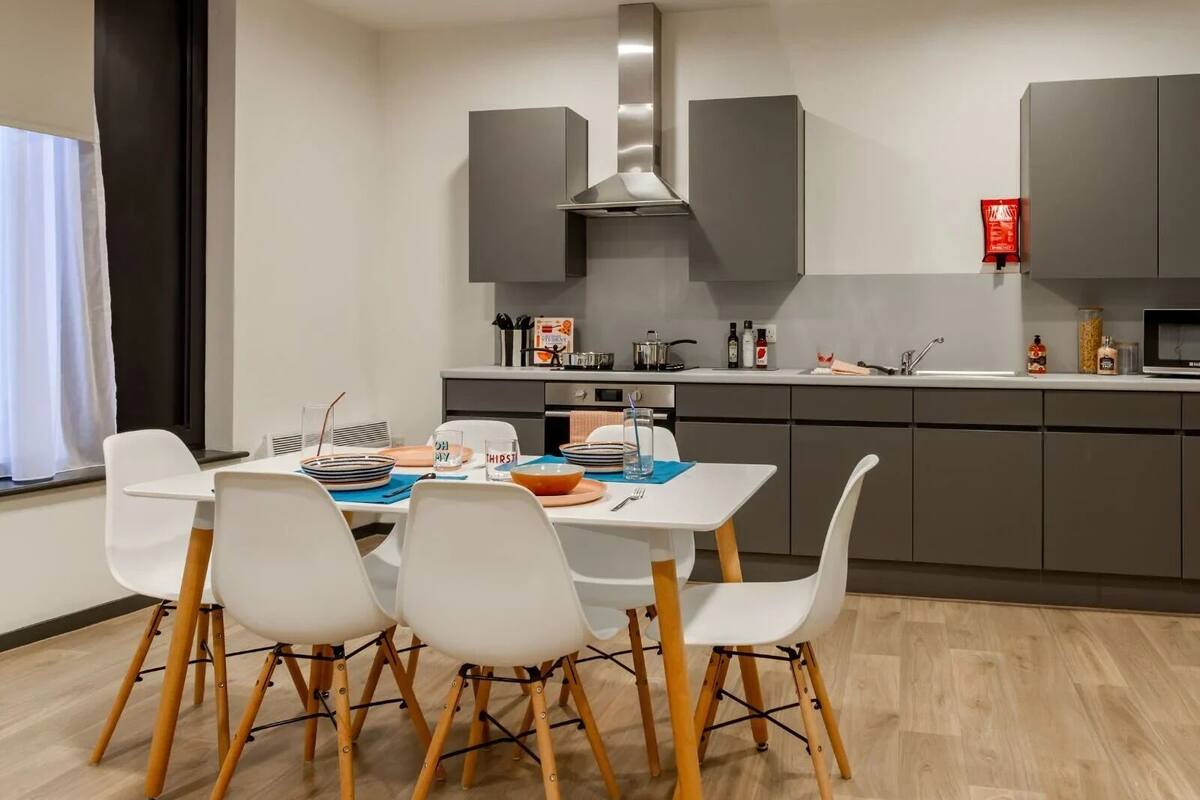 A modern kitchen with grey cabinets, a stainless steel range hood, and a dining area with white chairs and a wooden table.