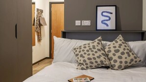 Una cama bien tendida con ropa de cama blanca y dos almohadas estampadas, una obra enmarcada sobre la cama, y una puerta con un abrigo colgado.