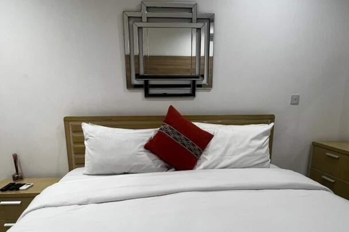 A neatly made bed with white bedding and a red accent pillow, a wooden headboard, a bedside table with a lamp, and a wall-mounted mirror.