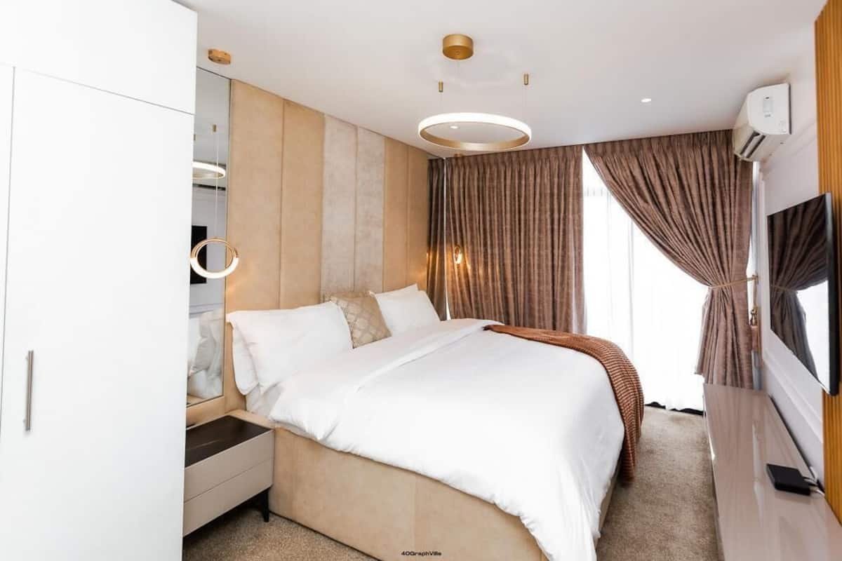 A modern hotel room with a large bed, a bedside table, a wardrobe, a TV, and a large window with curtains.