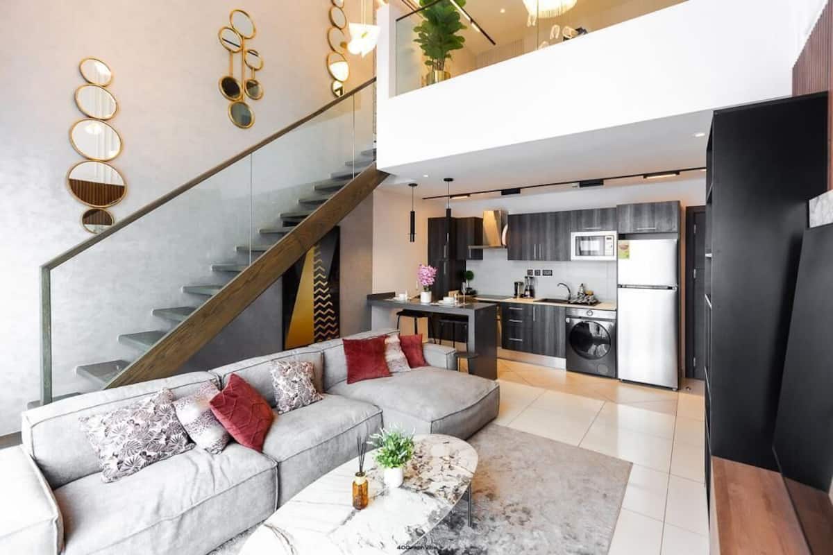 A modern apartment with a living area featuring a grey sectional sofa, a marble coffee table, and a staircase leading to an upper floor.