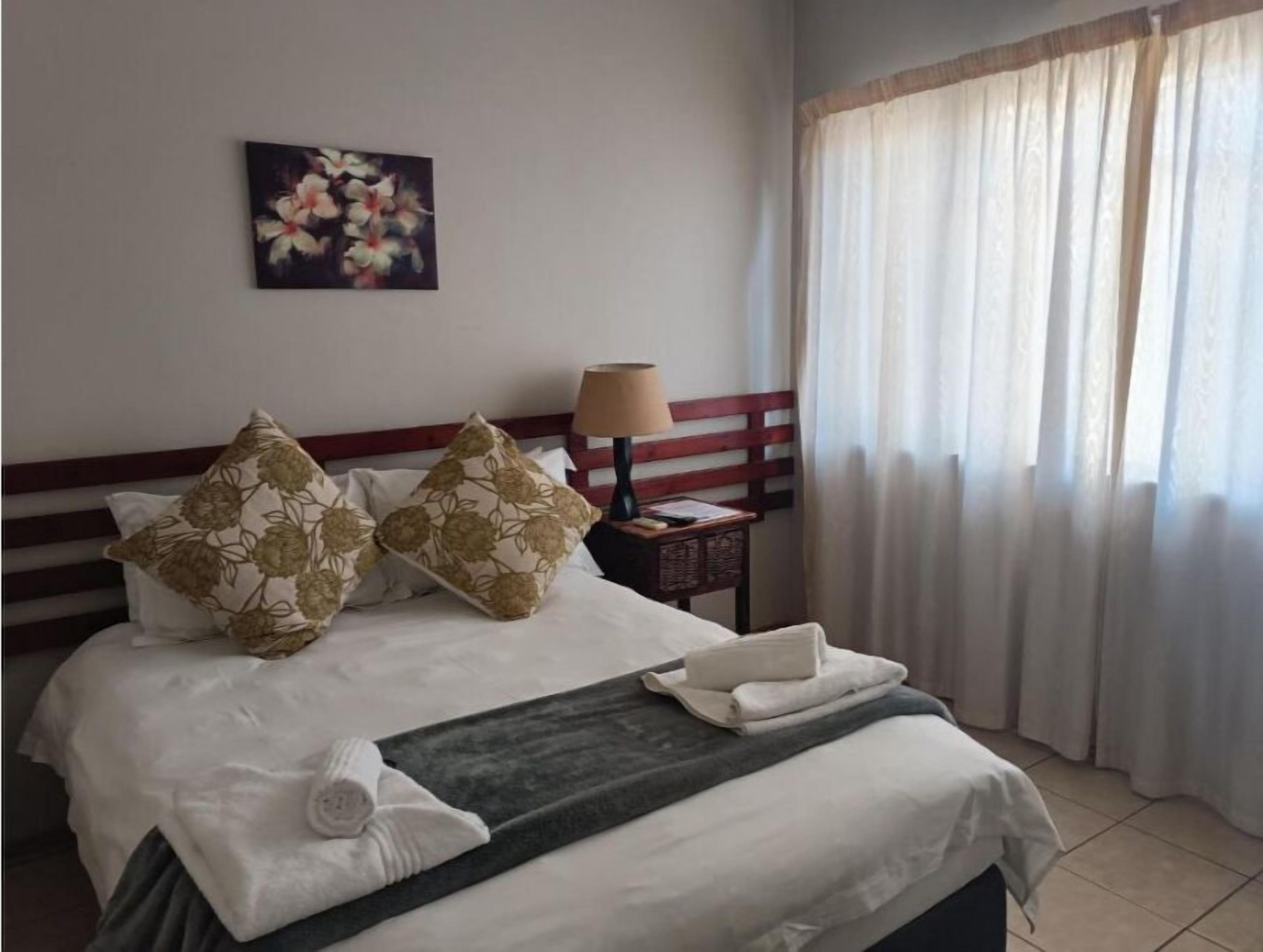 A hotel room with a bed, pillows, a bedside table with a lamp, a painting on the wall, and a window with curtains.
