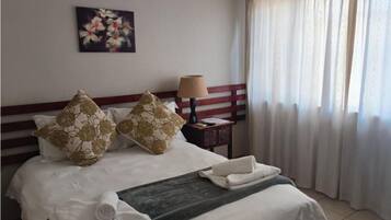 A hotel room with a bed, pillows, a bedside table with a lamp, a painting on the wall, and a window with curtains.