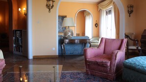 Un salon avec un fauteuil rose, un canapé bleu et un tapis à motifs. On y trouve une table basse en bois et une bibliothèque remplie de livres.