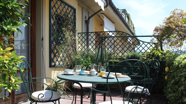 Une terrasse extérieure avec une table ronde et quatre chaises, entourée de verdure et d’un bâtiment doté d’une fenêtre à croisillons.