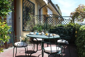 Een terras buiten met een ronde tafel en vier stoelen, omgeven door groen en een gebouw met een lattenraam.
