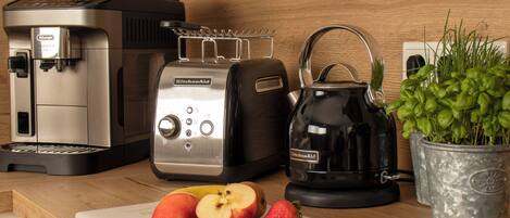 A kitchen counter with a coffee maker, toaster, kettle, and a cutting board with apples and strawberries.