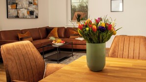 A modern living room with a brown leather sofa, a wooden coffee table, and a dining table with chairs. A vase of colorful flowers is on the coffee table. - Idyllic vacation apartment 300m from the Zwischenahner Meer (Bad Zwischenahn)