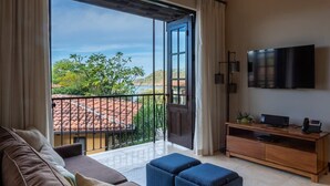 A modern living room with a sofa, ottomans, a TV, and a balcony with a view of greenery and a tiled roof. - Beautiful Mediterranean-Style Penthouse with an Oceanview Balcony (Las Catalinas)