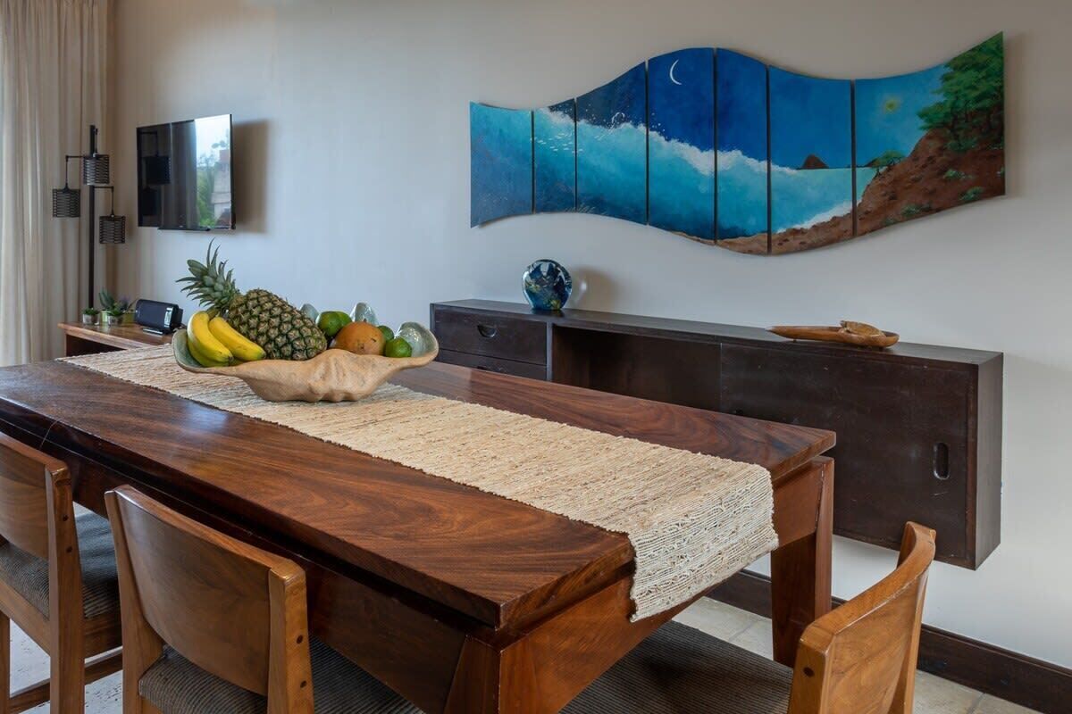Un coin repas avec une table en bois, des chaises, un set de table en fibres tressées et un panier de fruits. Un téléviseur à écran plat est fixé au mur au-dessus de la table.