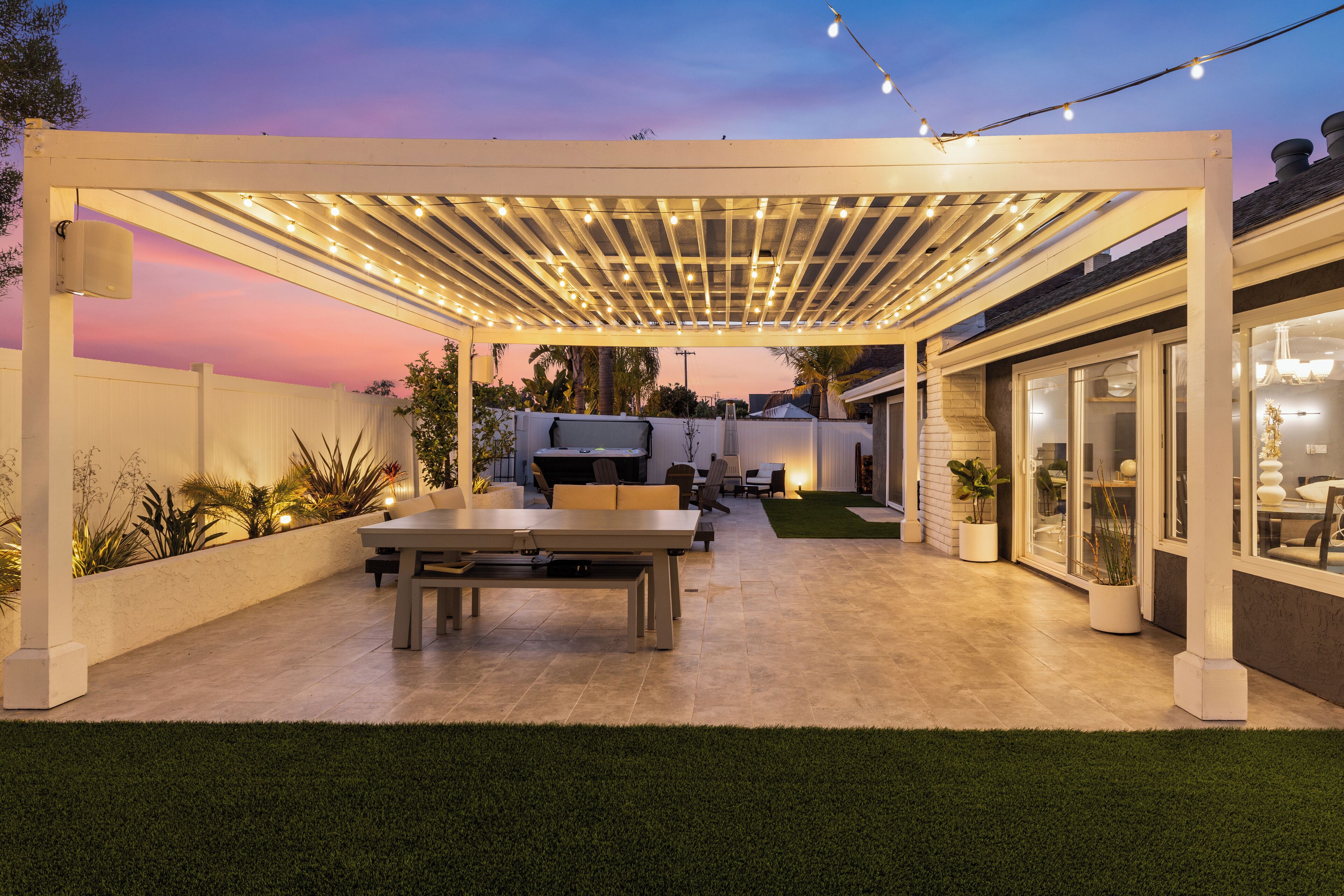 A covered patio area with a dining table and chairs, surrounded by potted plants and a white fence.