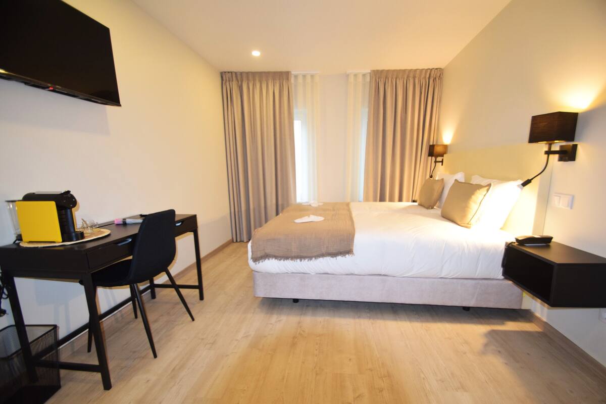 A hotel room with a bed, a desk, a chair, a TV mounted on the wall, and a window with curtains.