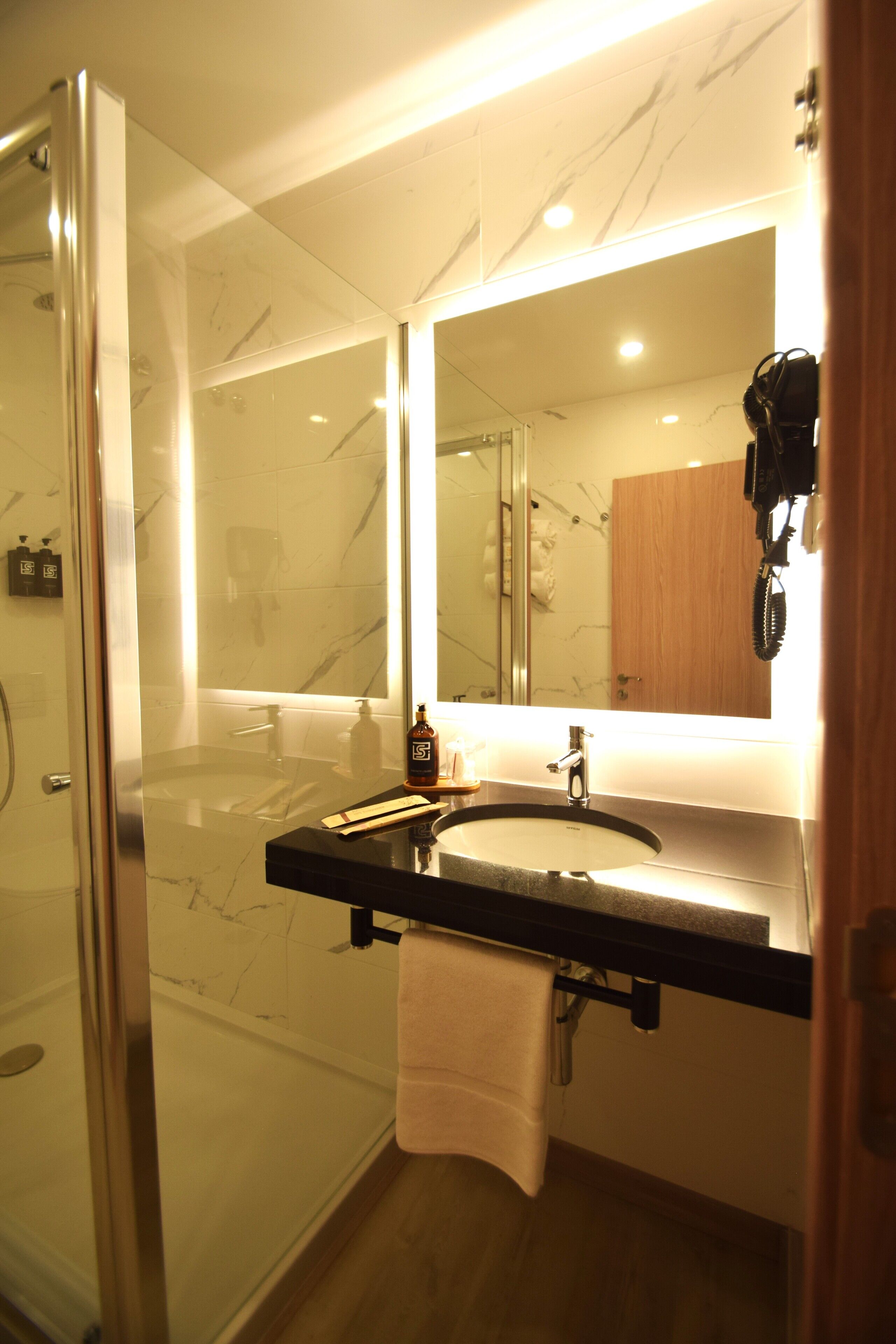 A modern bathroom with a glass-enclosed shower, a marble-tiled wall, a black countertop, and a large mirror.