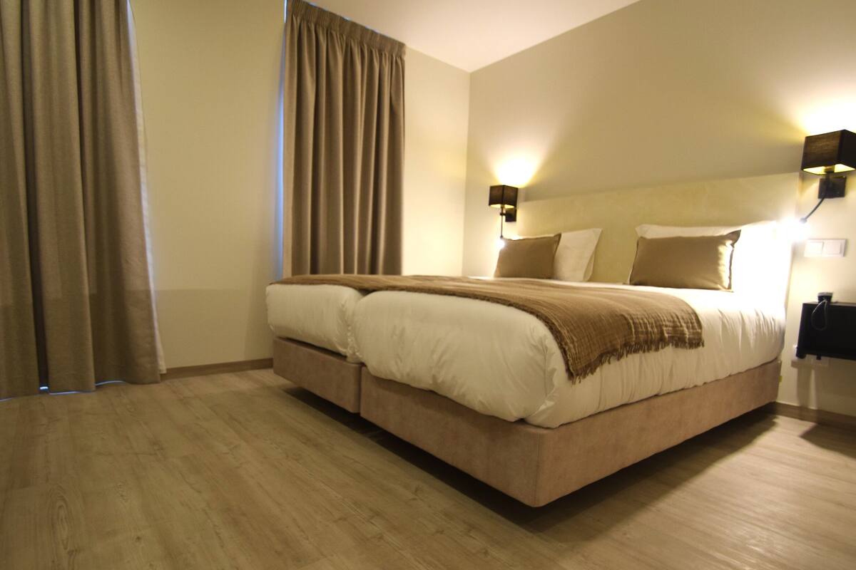 A hotel room with a large bed, two bedside tables, a wall-mounted lamp, and a curtain.