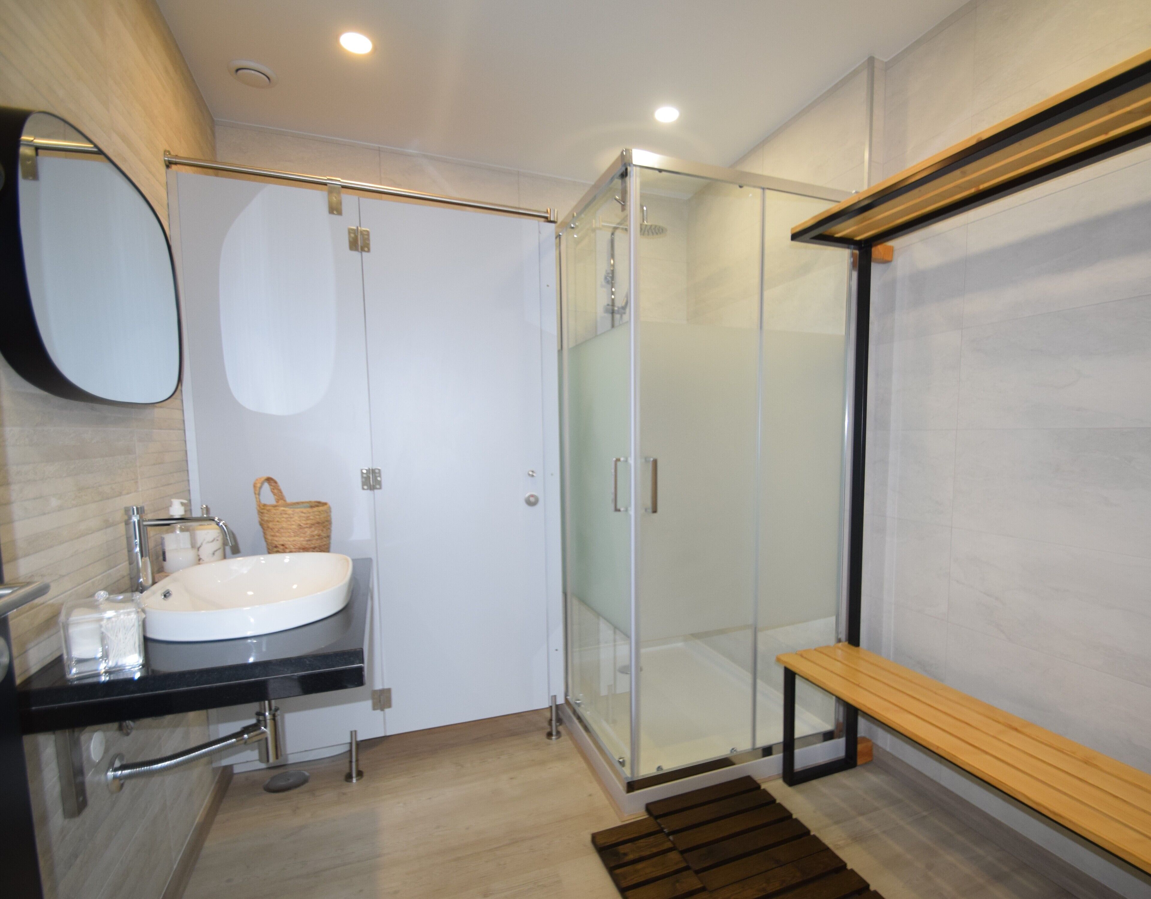 A modern bathroom with a glass shower enclosure, a wooden bench, and a white sink.