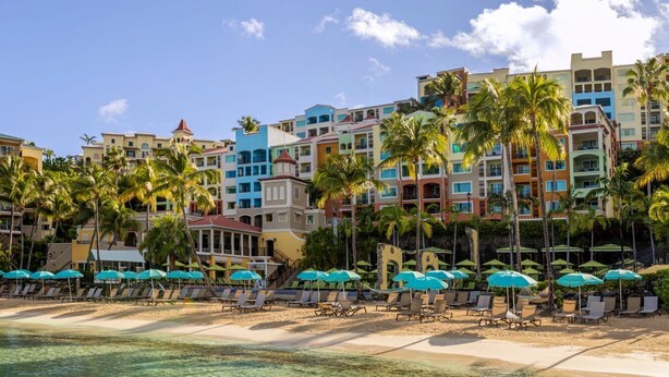 Un hôtel situé en bord de mer, composé de plusieurs bâtiments aux couleurs vives, avec une plage de sable, des chaises longues et des parasols, ainsi que des palmiers.