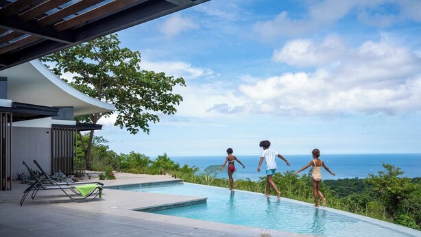A swimming pool with a view of the ocean, surrounded by greenery and modern architecture. - Luxurious Ultramodern Property with Private Suites, Infinity Pool, and Sweeping Ocean and Mountain Views (Sardinal)