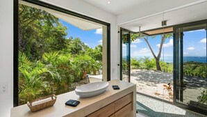 A modern bathroom with a large window, a white sink, and a view of a lush garden and ocean. - Luxurious Ultramodern Property with Private Suites, Infinity Pool, and Sweeping Ocean and Mountain Views (Sardinal)