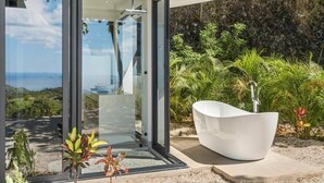 A modern house with a glass door leading to a patio with a white bathtub and a view of the ocean. - Luxurious Ultramodern Property with Private Suites, Infinity Pool, and Sweeping Ocean and Mountain Views (Sardinal)