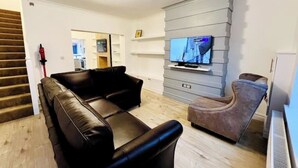 A living room with a black leather sofa, a brown armchair, a flat-screen TV mounted on a grey wall, and a staircase in the background. - Modern 2-bed house  in Blyth centre  with two bedrooms and two bathrooms (Blyth)