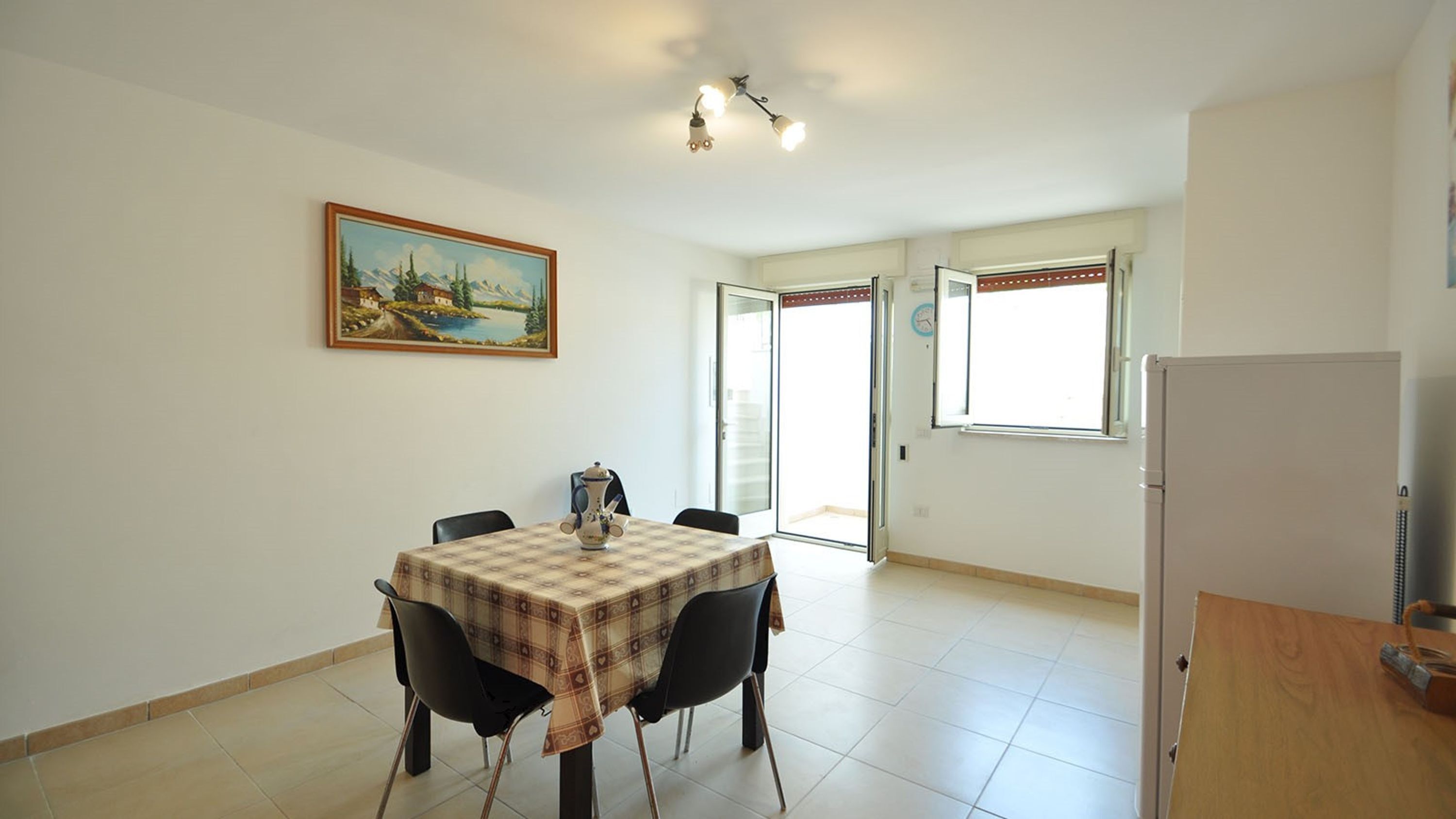 A dining area with a table set for two, a painting on the wall, and a refrigerator in the corner.