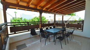 An outdoor patio with a wooden ceiling, a table with chairs, and a view of greenery and buildings. - Villa Romano 9 Guests Torre Suda (Racale)