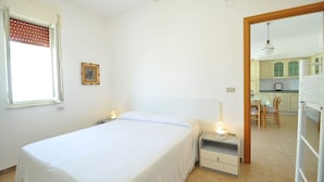 Una camera da letto con un letto, comodini, una lampada, una finestra con persiane e una porta che dà su una cucina.