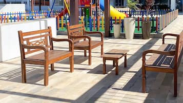 Un espacio al aire libre con techo, con bancos y mesas de madera, una estructura de juegos para niños al fondo y vista a edificios residenciales.