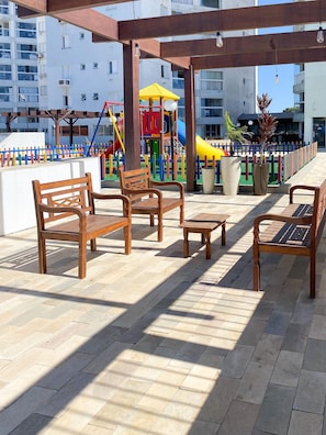 A covered outdoor seating area with wooden benches and tables, a children's play structure in the background, and a view of residential buildings. - Resid. Clube Mar Grosso-Laguna - LAG706 (Laguna)