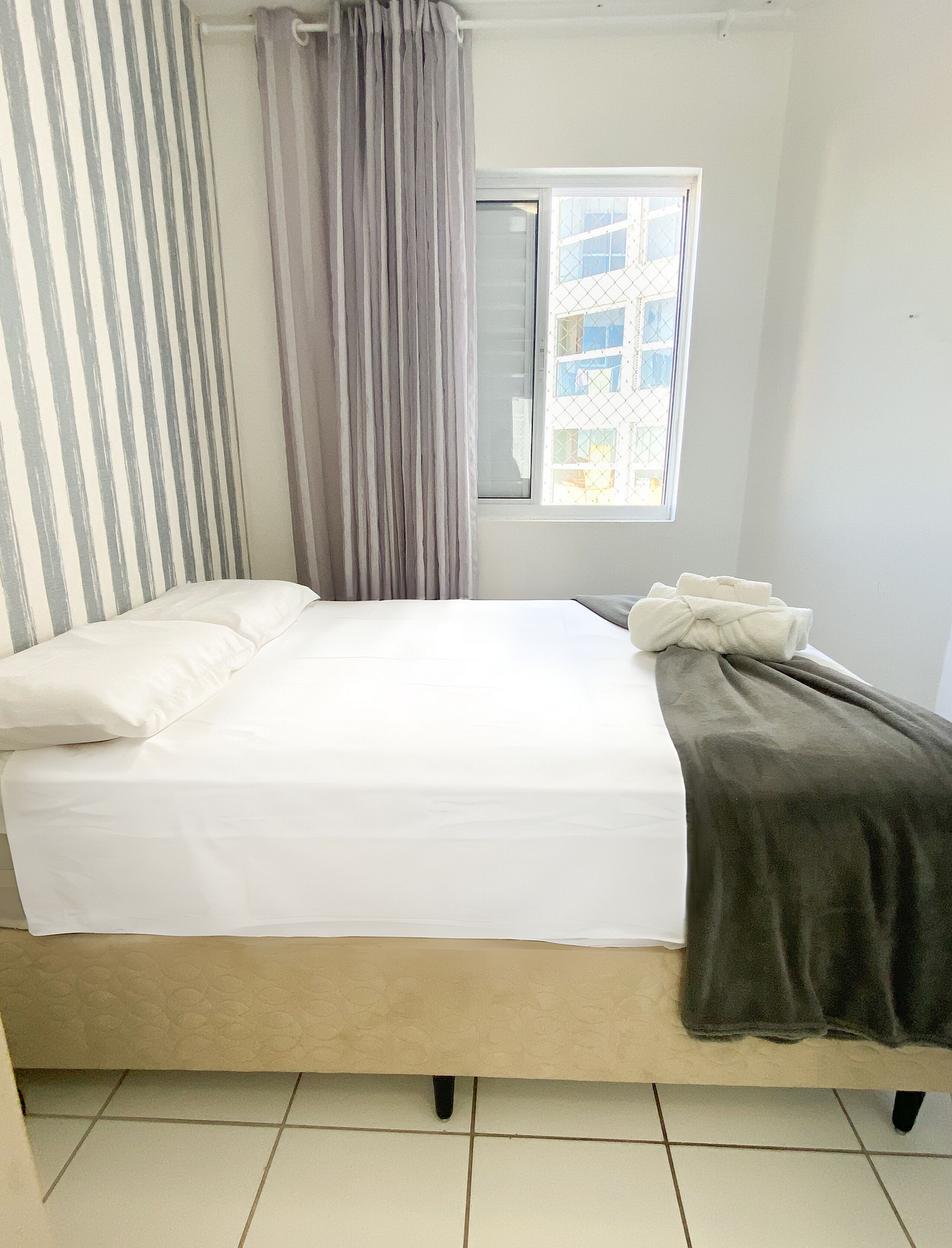 Uma cama com colchão branco, cobertor cinza e cortina listrada.