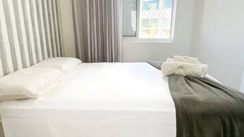 Uma cama com colchão branco, cobertor cinza e cortina listrada.