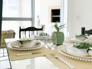 Una mesa de comedor dispuesta con platos blancos, vasos transparentes y un jarrón verde con una planta.