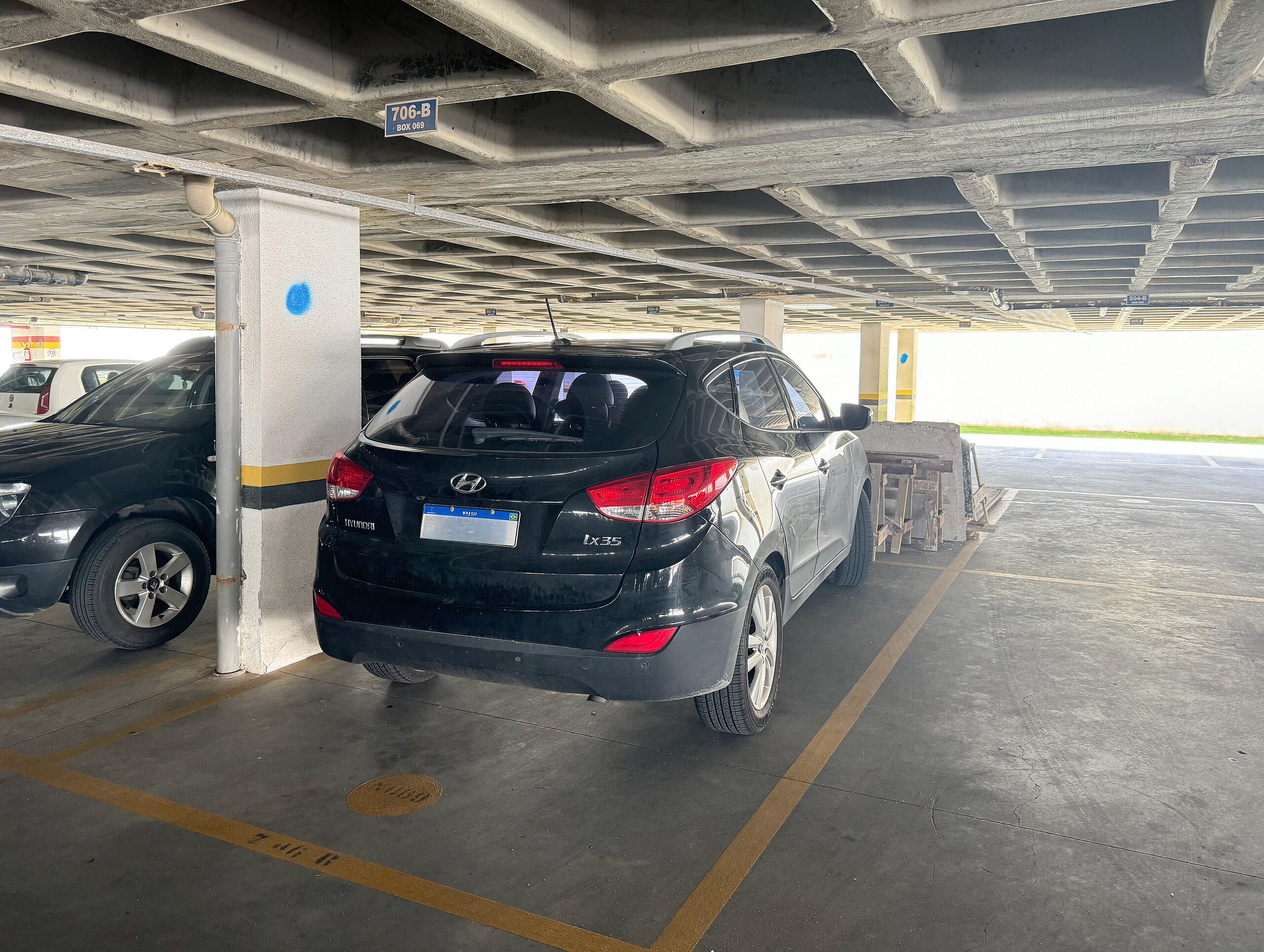 Un estacionamiento con una camioneta SUV negra estacionada cerca de un pilar, techo de concreto y un letrero que indica '706-B BOX 069'.
