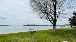 A grassy area with a dock extending into a lake, a large tree, and a distant shoreline with buildings. - Comfortable 3-bedroom lake house with AC, WiFi on Devils Lake in Addison, MI. (Addison)