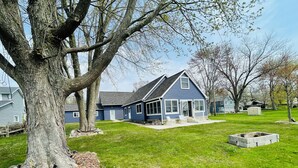 A blue house with a porch, a stone fire pit, and a large tree in the foreground. - Comfortable 3-bedroom lake house with AC, WiFi on Devils Lake in Addison, MI. (Addison)