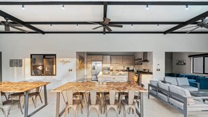 A spacious living area with a large wooden dining table, a sofa, and a kitchen in the background. - Brand-New 6BD Giant with Private Pool by SPCR (Tempate)