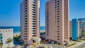 Un hôtel de grande hauteur, avec de nombreux étages, offrant une vue sur la plage et disposant d’un parking pouvant accueillir plusieurs véhicules.