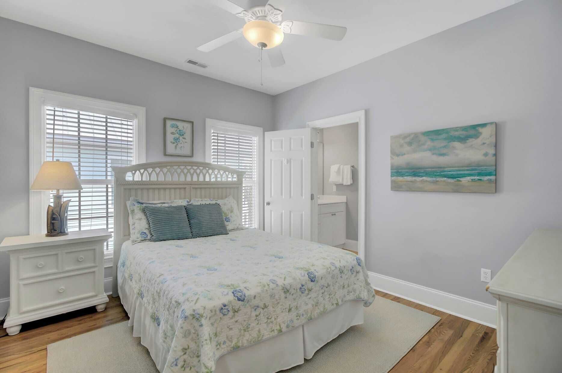 A bedroom with a bed, a nightstand, a dresser, a ceiling fan, a painting, and a door leading to another room.