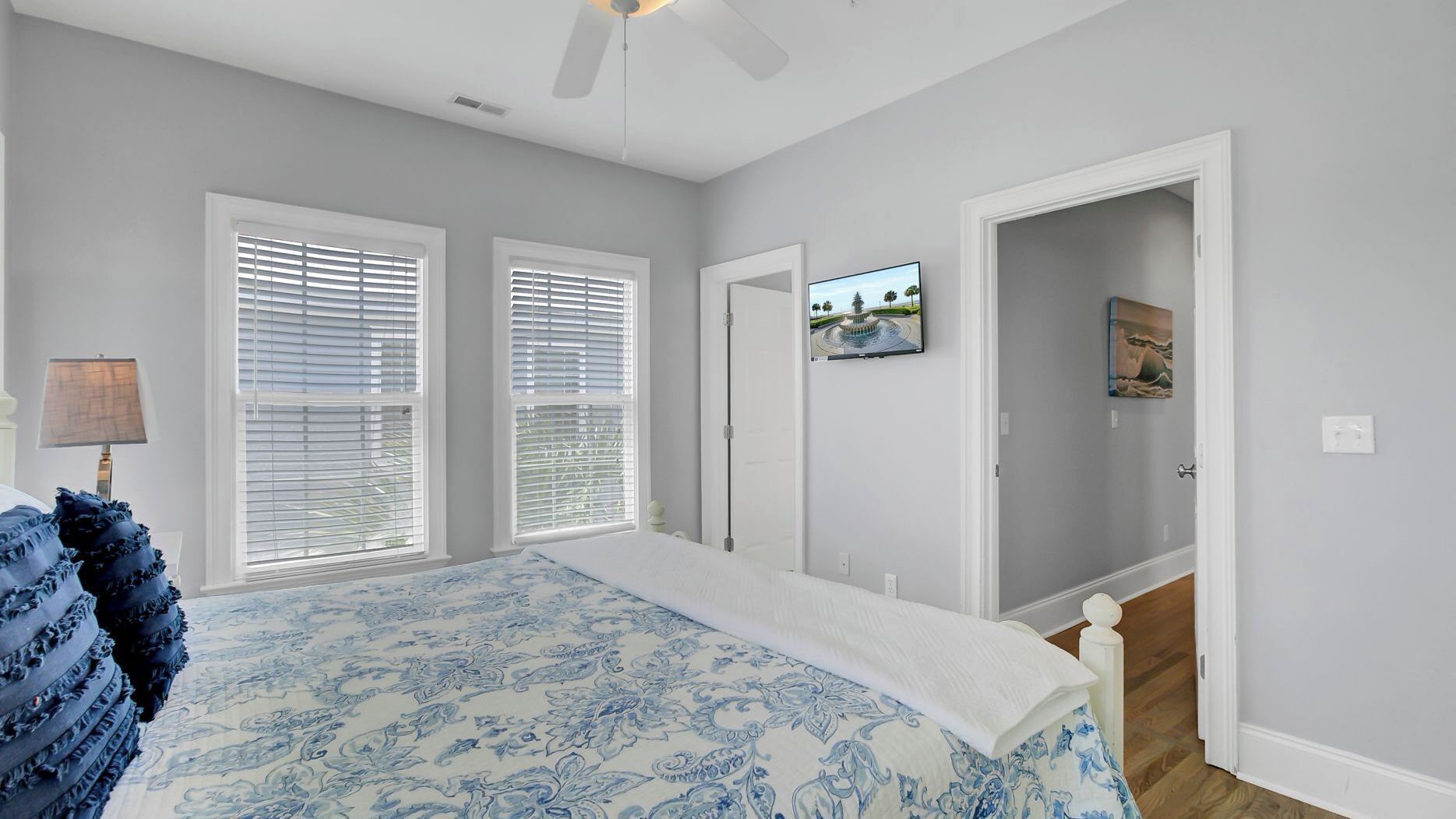 A bedroom with a bed, two windows with blinds, a ceiling fan, a TV, and a door leading to another room.