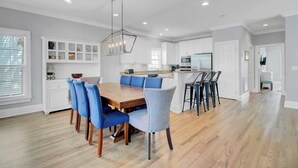 A modern kitchen with white cabinets, a wooden dining table, blue chairs, and a pendant light. - 6 10th Avenue by Avantstay Steps to the Beach Pool 2691 Sqft (Isle of Palms)