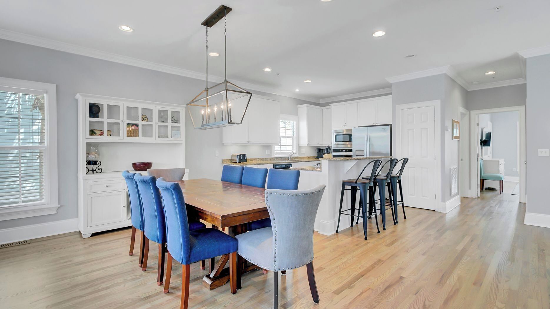 A modern kitchen with white cabinetry, a wooden dining table with blue upholstered chairs, and a hanging light fixture.