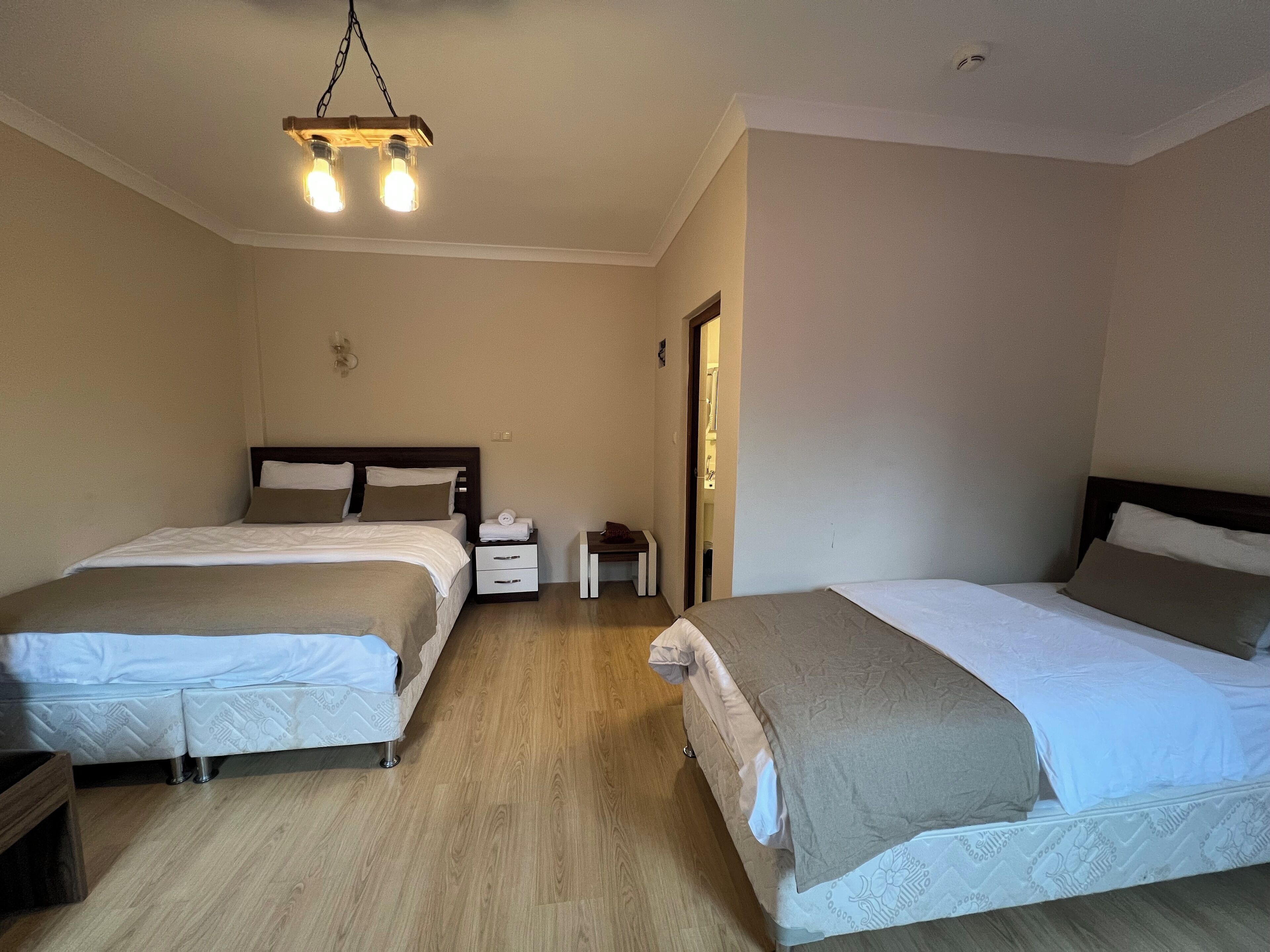 A hotel room with two beds, wooden flooring, a small bedside table, and a hanging light fixture.