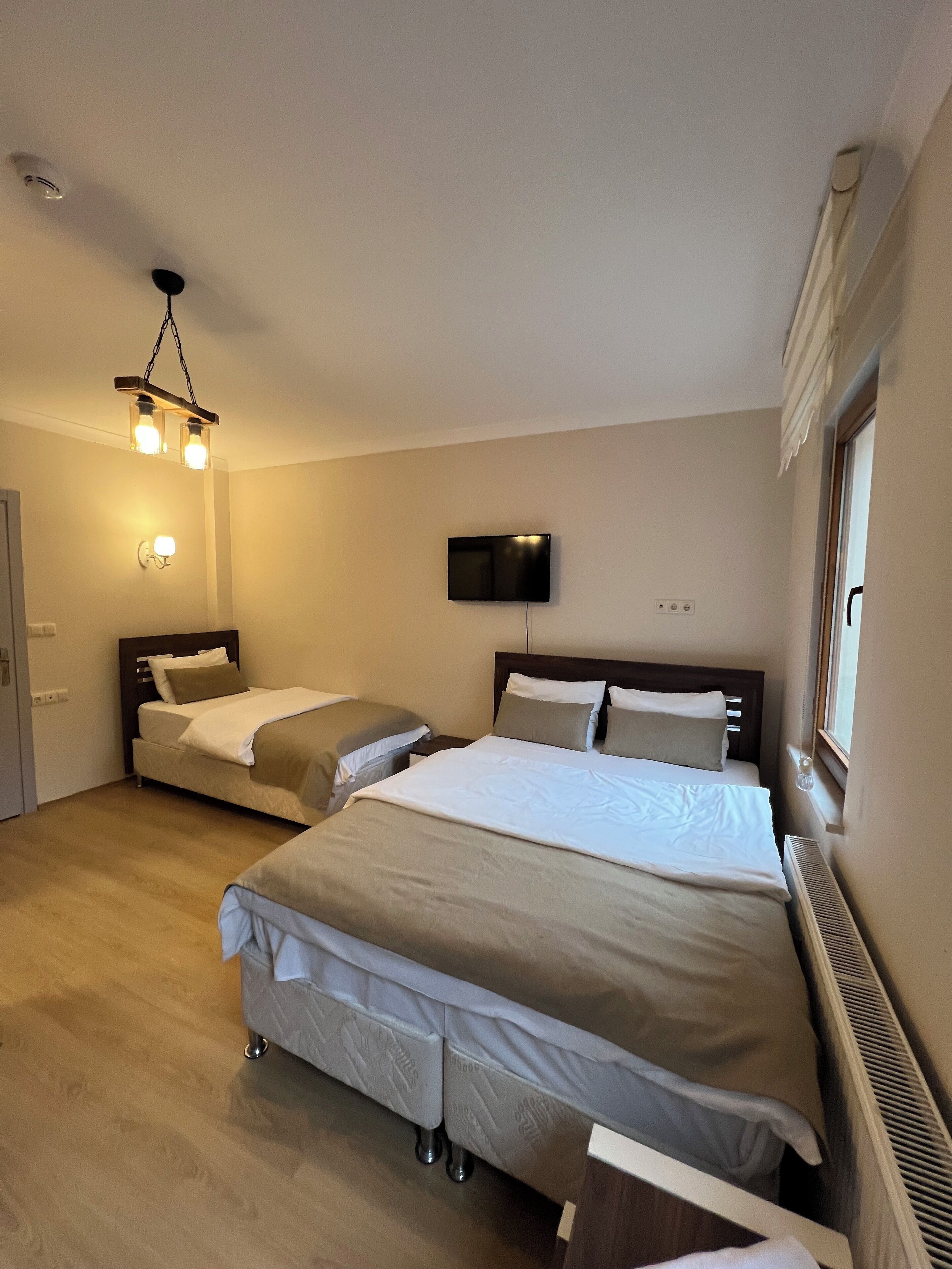 A hotel room with two beds, a flat-screen TV mounted on the wall, a ceiling light fixture, and a window with a view.