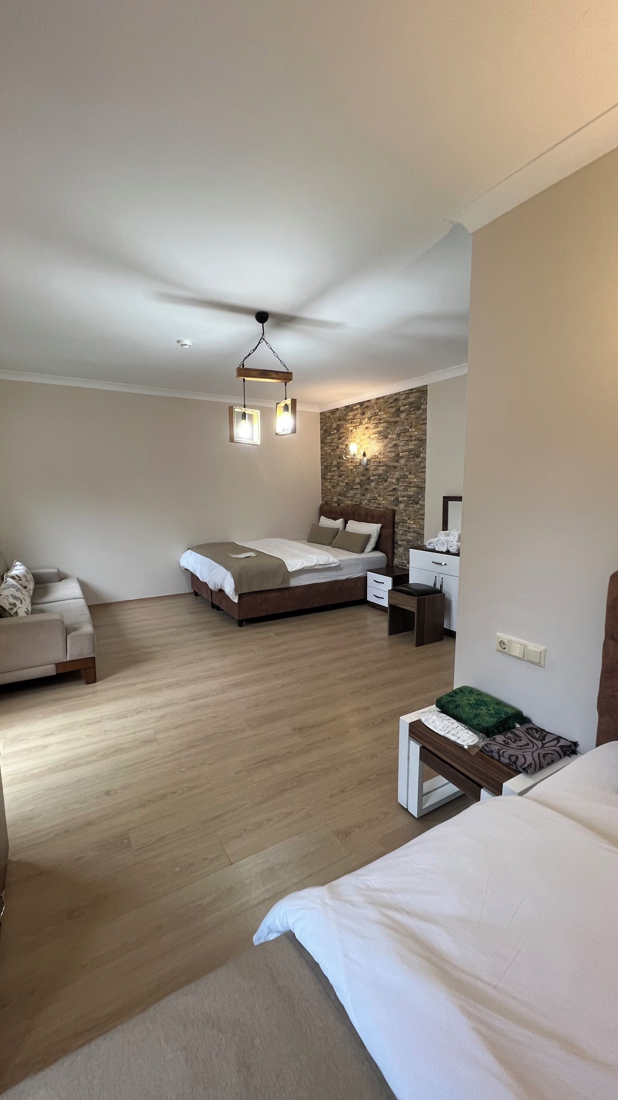 A modern hotel room with a large bed, a sofa, a desk, and a stone accent wall.