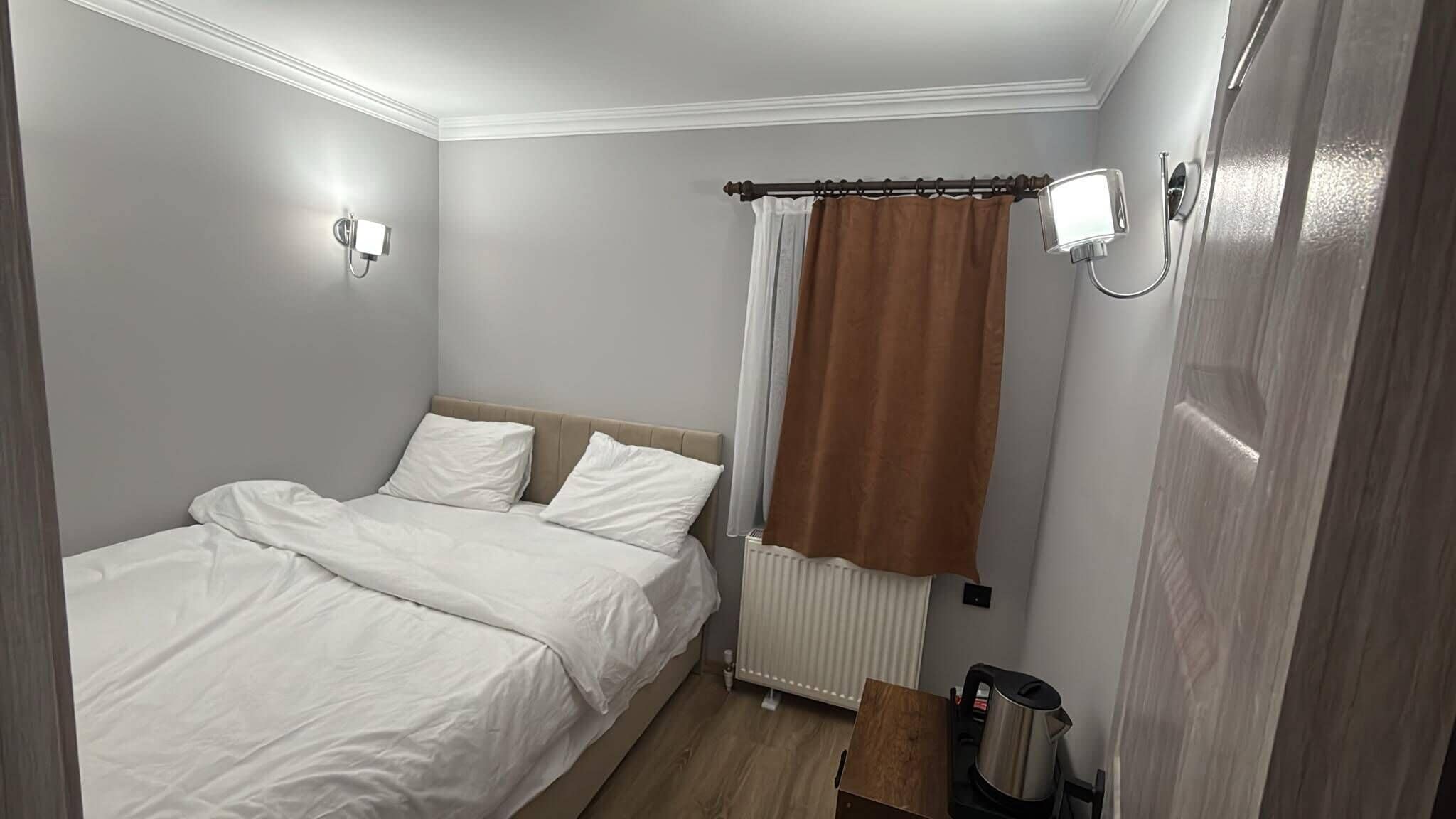 A hotel room with a bed, a radiator, a kettle, and a curtain.