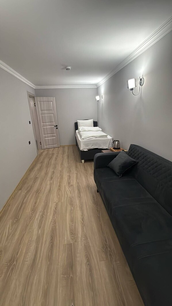 Une pièce comprenant un lit, un canapé noir, un plancher en bois, une porte et un luminaire fixé au mur.