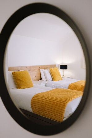 A hotel room with a wooden headboard, two beds with white bedding and yellow accents, a black lamp on a bedside table, and a mirror reflecting the room. - The Old Farmhouse (Truro)
