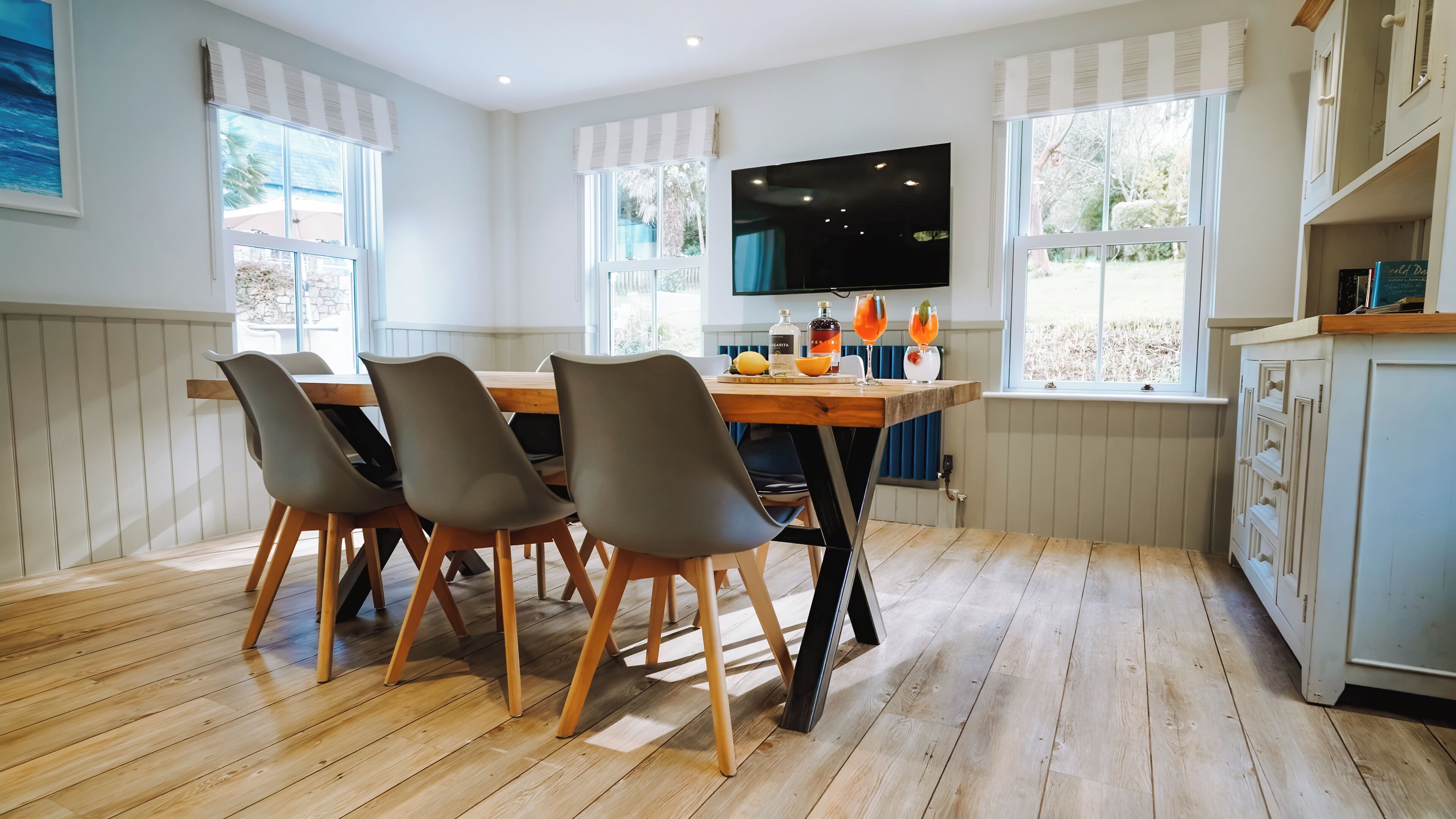 A dining area with a wooden table, four chairs, a TV mounted on the wall, and windows with blinds.