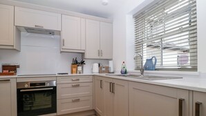 A modern kitchen with white cabinets, a built-in oven, a sink, and a window with blinds. - Capella Cottage (Keswick)